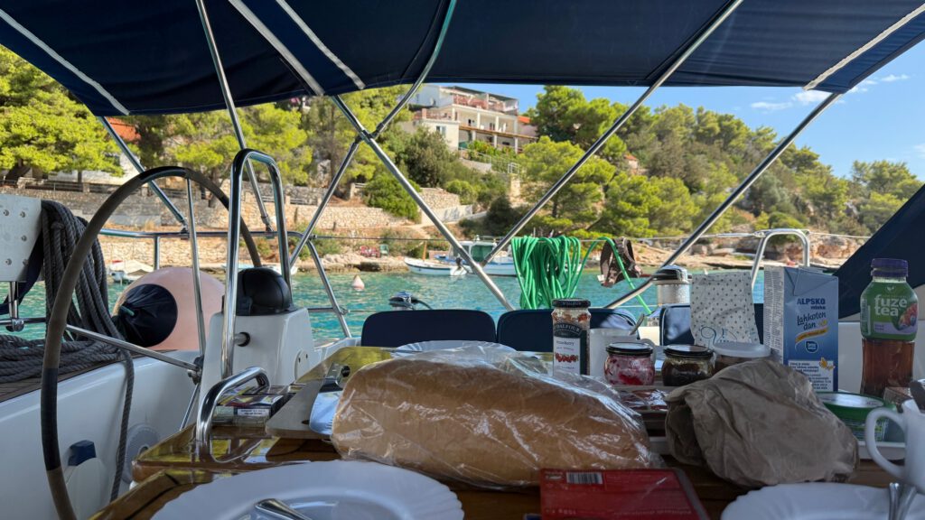 Gedeckter Frühstückstisch im Cockpit mit Brot, Marmelade und Saft, dahinter eine Bucht mit Pinien