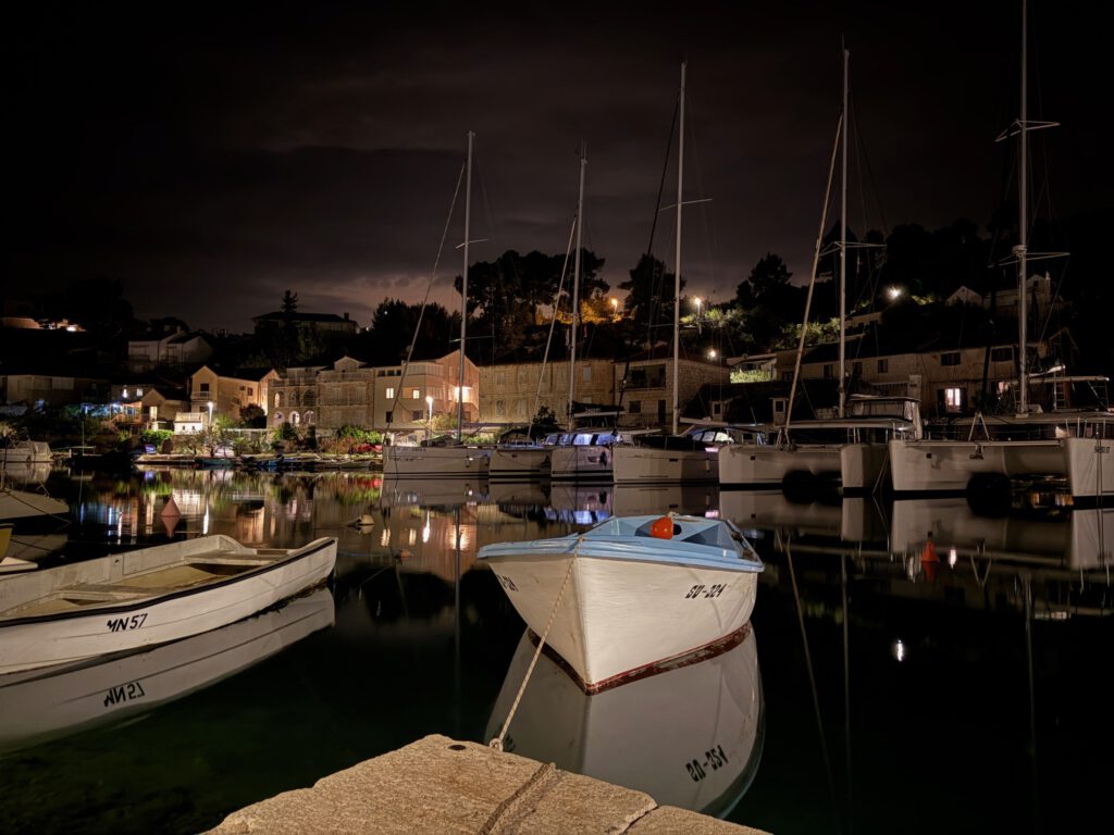 Kleines weißes Boot an der Hafenkante, dahinter beleuchtete Häuser und Segelyachten in der Marina von Bobovišća auf Brač bei Nacht